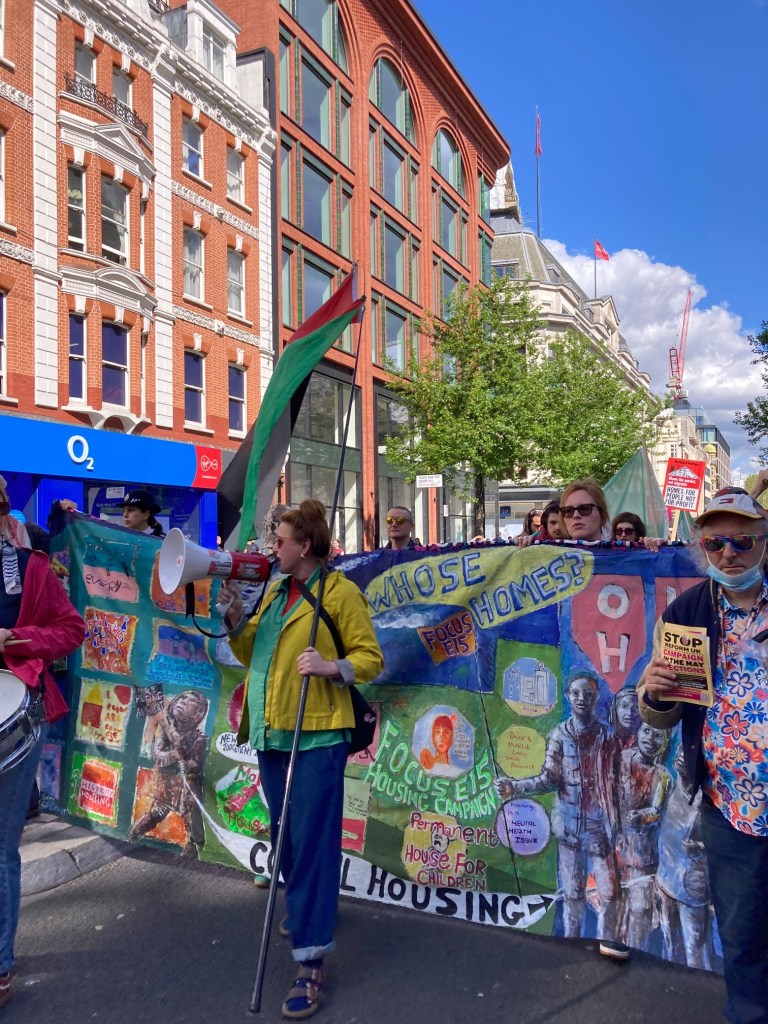 Photo of a white woman wearing jeans and a yellow jacket holding a megaphone in one hand and a Palestinian flag in the other while walking in front of a large multi-coloured banner held up by fellow campaigners that includes phrases such as ‘Whose Homes?’, ‘Focus E15 housing campaign’ and ‘Permanent house for children’.