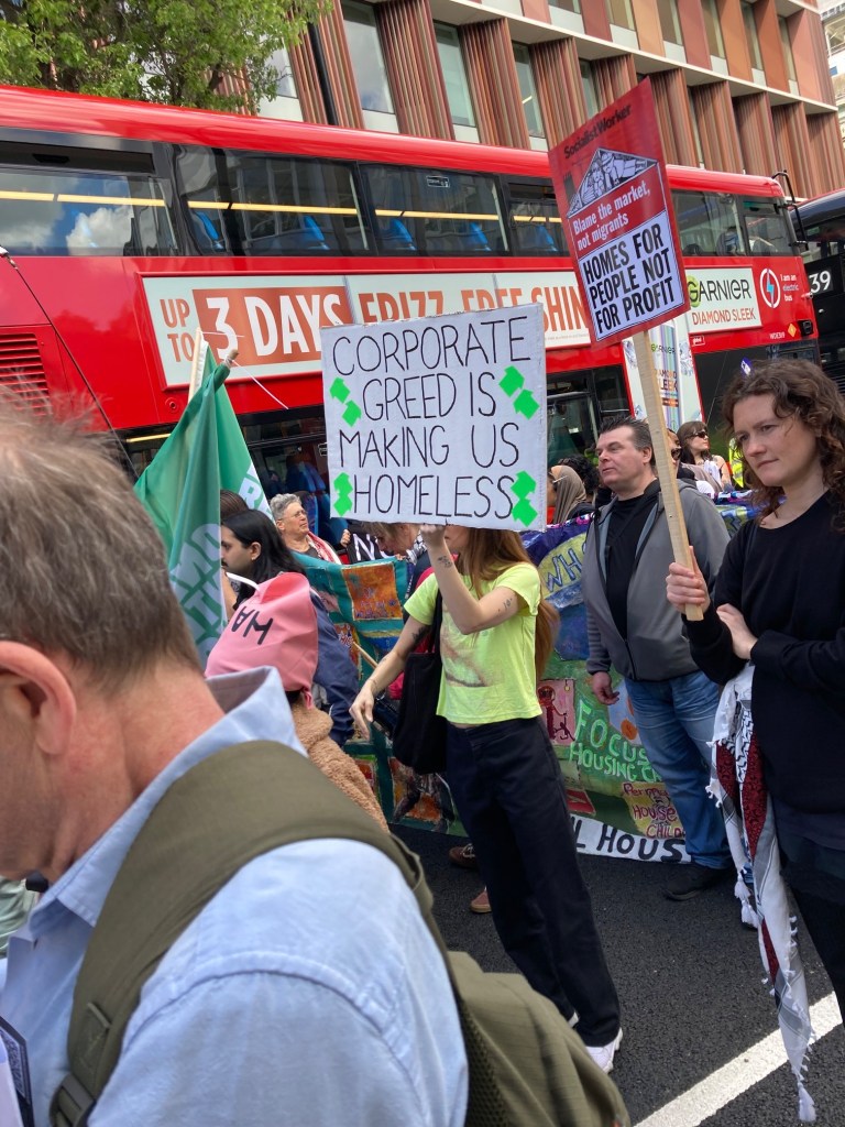 Picture of a person in a crowd of protesters holding up a white home-made sign that reads ‘CORPORATE GREED IS MAKING US HOMELESS’ in black letters. A double-decker bus is visible in the background, and the person walking in front is wearing a pink ‘HASL’ scarf around their head.