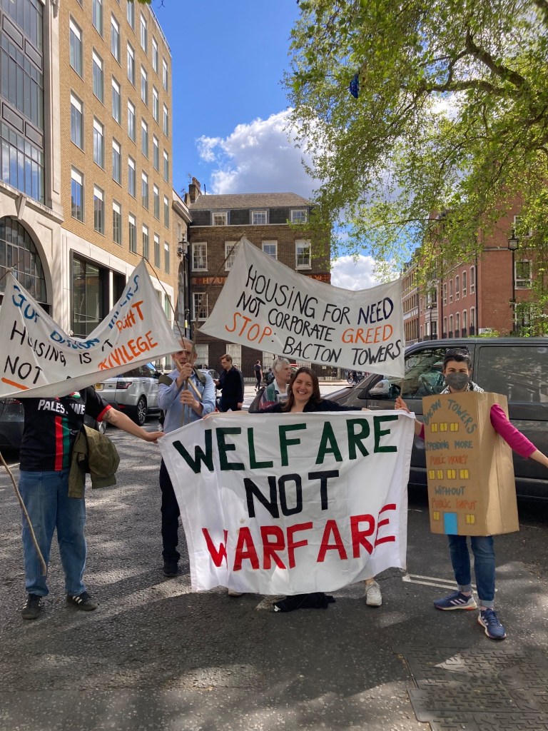 Members of BTA and Camden Friends of Palestine holding up signs that read ‘WELFARE NOT WARFARE’ AND ‘HOUSING FOR NEED NOT CORPORATE GREED - STOP BACTON TOWERS’. One BTA member is wearing a cardboard box with windows and doors painted on it that reads ‘Bacton Towers - spending more public money without public input’.