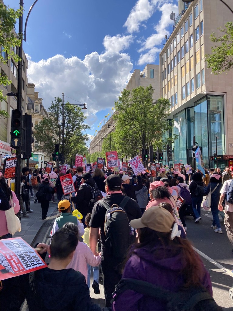 Photo of campaigners holding placards that read ‘Homes for people not profit’ are walking down Oxford Street in the sunshine, with a blue sky visible above. 